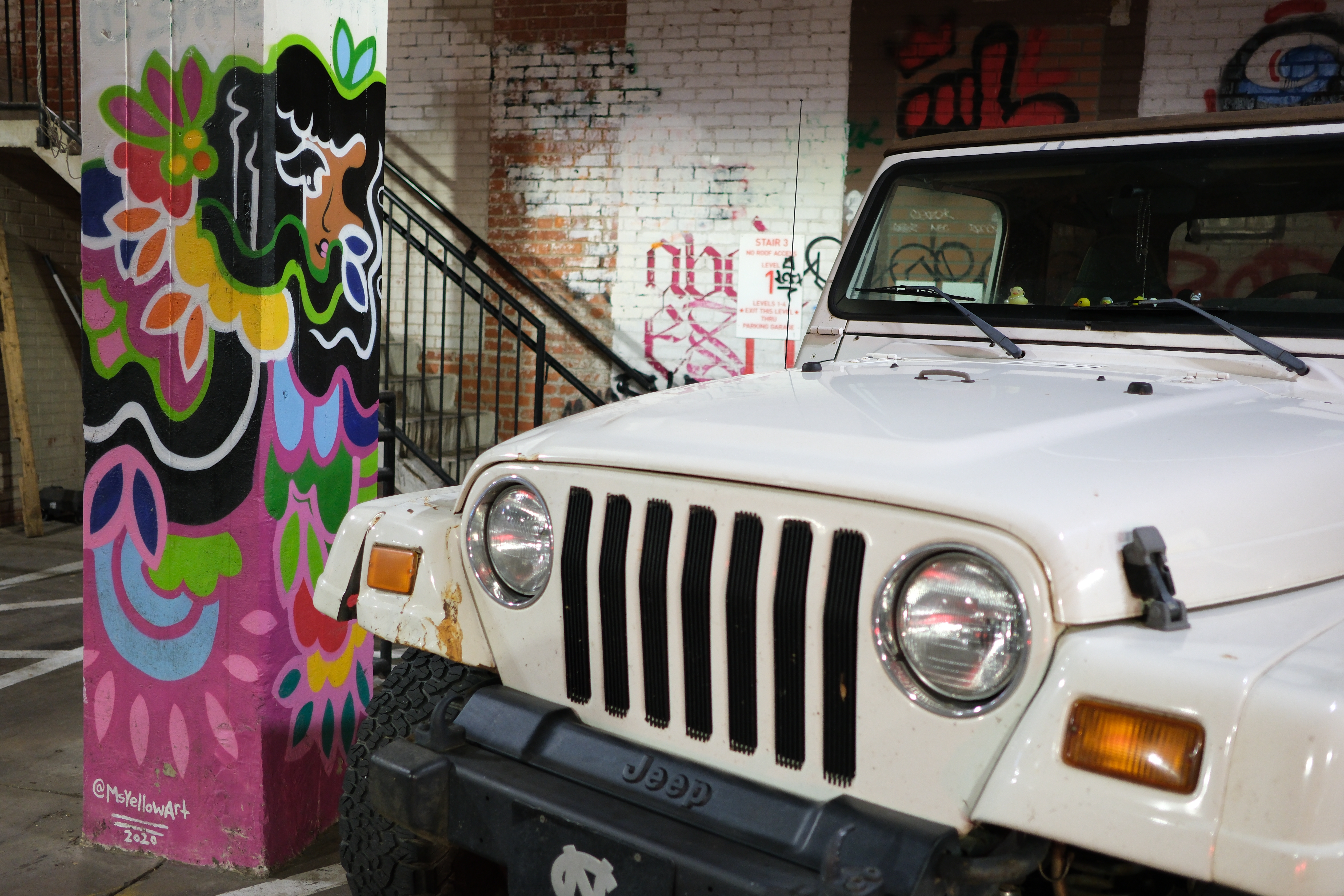 Familiar Faces - a colorful mural of a woman, with her hair flowing and flowers enveloping her face, on a pillar in a parking lot. next to the mural a Jeep is parked. it is rusted above the passenger side front wheel. there are six small rubber ducks on the dash.
