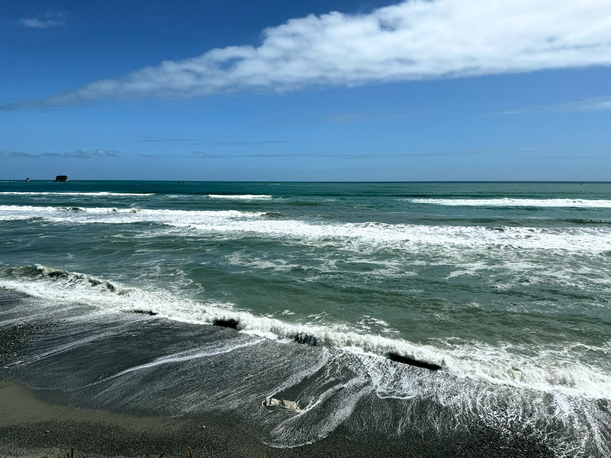 The coast alongside the Tasman Sea. The sky is blue with some cloud cover. The waves find their way to the shore from afar. The water is receding at the time the photo was taken.